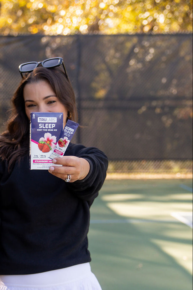 Woman holding a TruLabs Sleep Strawberry Hibiscus product package on a tennis court